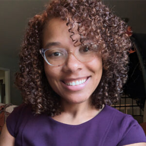 Carmen Seantel, smiling in a portrait photo, wearing glasses and a purple top, with curly hair and a cozy background.